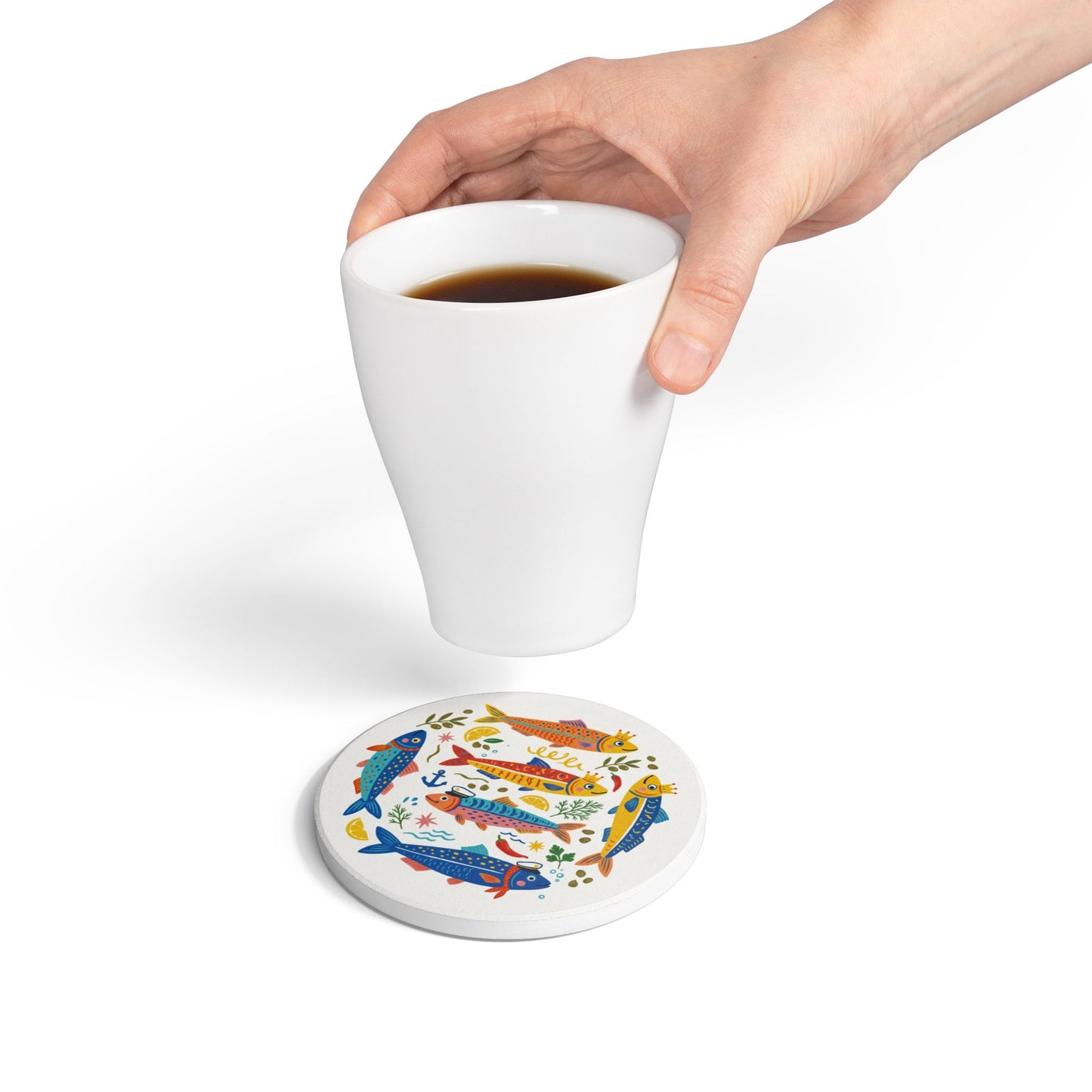 A hand placing a white coffee mug onto the colorful sardine ceramic coaster to demonstrate size and lifestyle use.