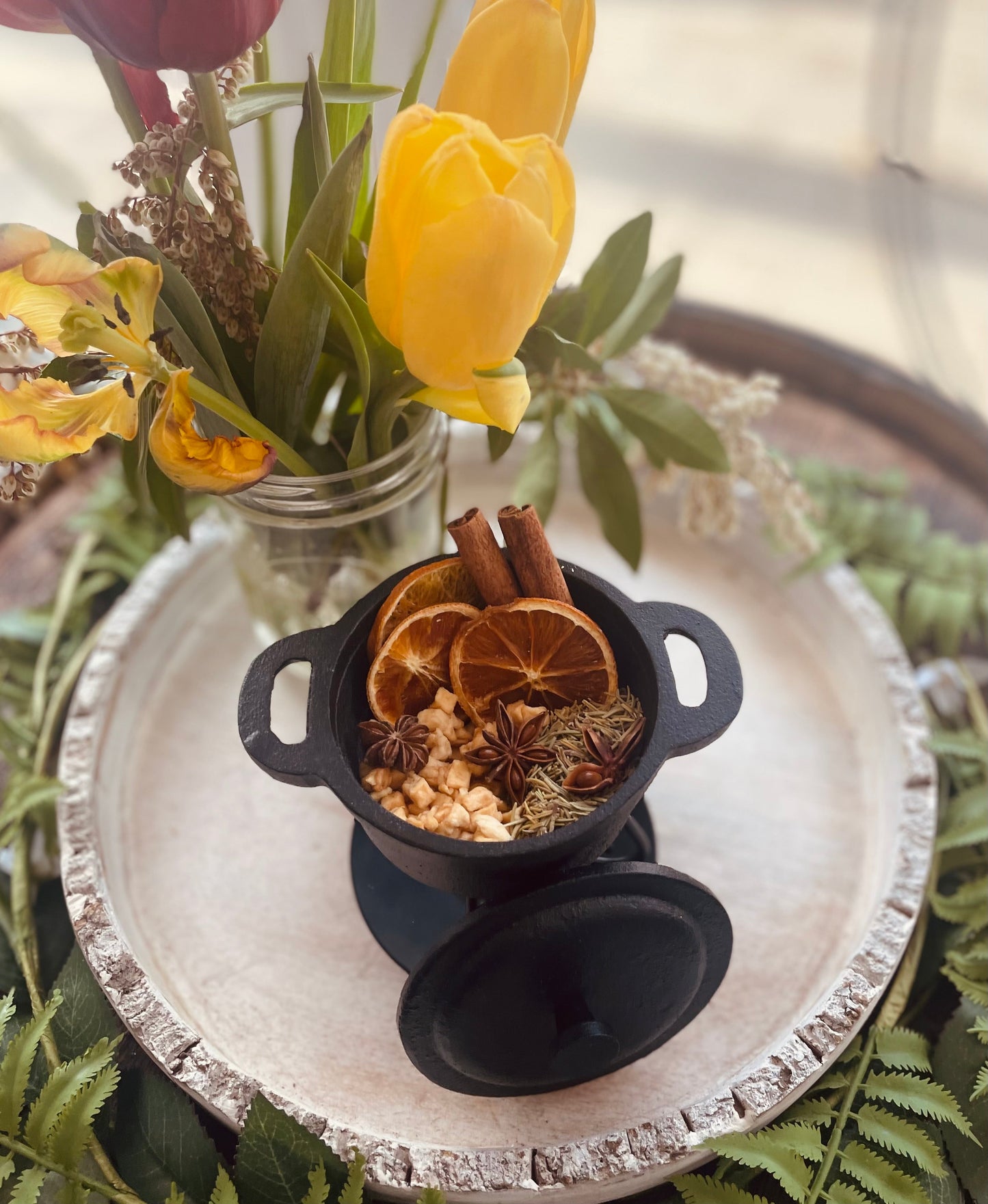 A close-up lifestyle shot of a prepared natural simmer pot blend on a rustic tray with vibrant yellow spring tulips softly blurred in the background.