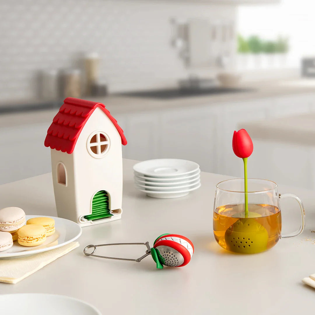 Red and green tulip tea infuser resting on a kitchen counter next to a decorative white ceramic house.