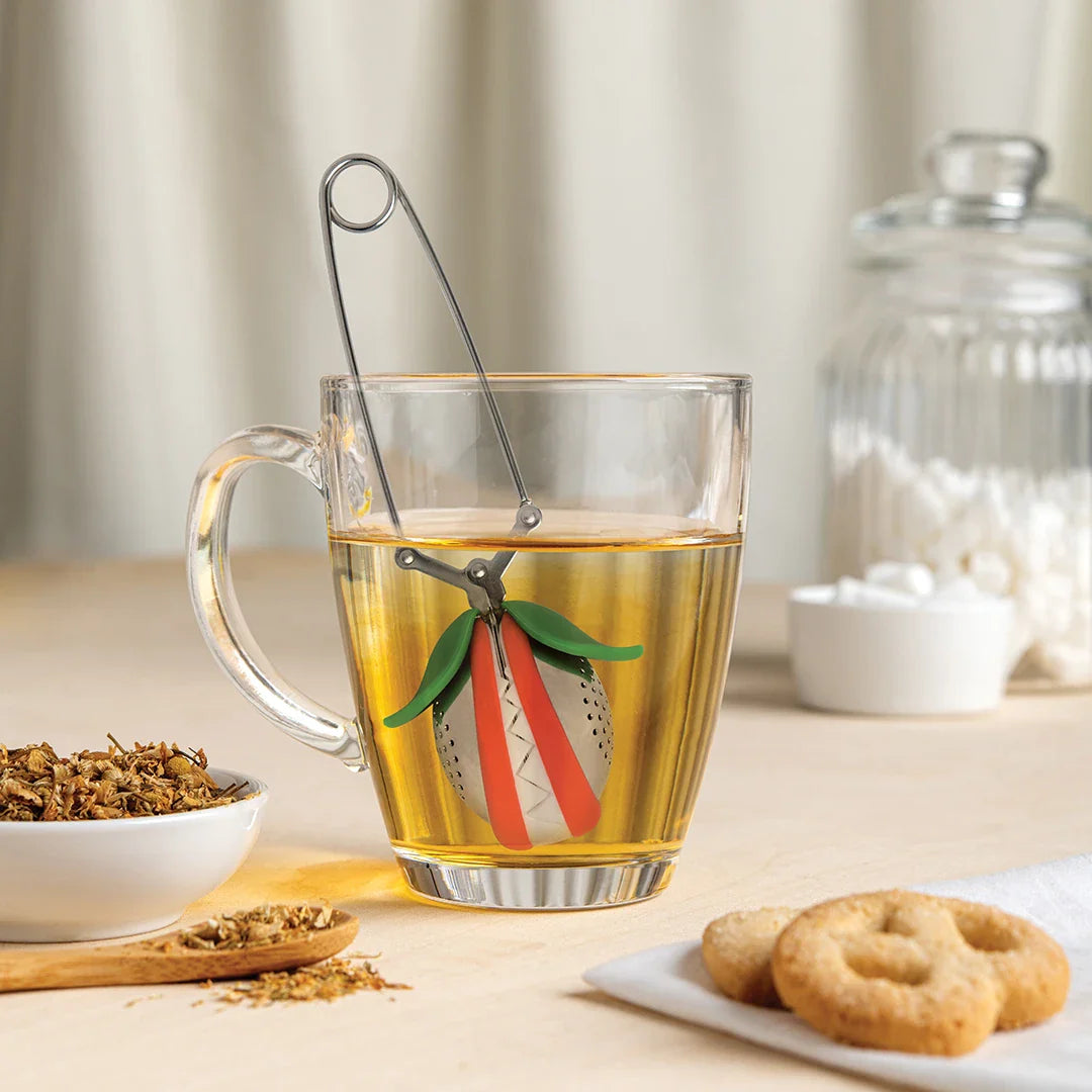 Tulip tea infuser sitting inside a clear glass mug of steeping amber tea next to biscuits on a kitchen counter.