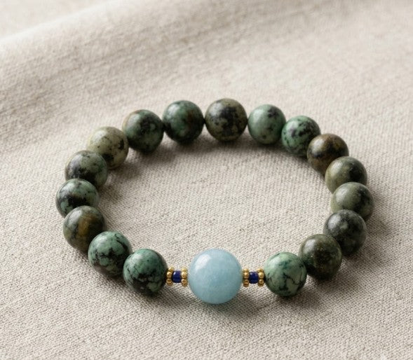 A close-up flatlay shot of a round gemstone stretch bracelet made of green African turquoise beads, tiny blue lapis spacers, gold-plated accents, and a central light blue quartz bead on a textured linen background.