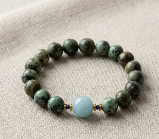 A close-up flatlay shot of a round gemstone stretch bracelet made of green African turquoise beads, tiny blue lapis spacers, gold-plated accents, and a central light blue quartz bead on a textured linen background.