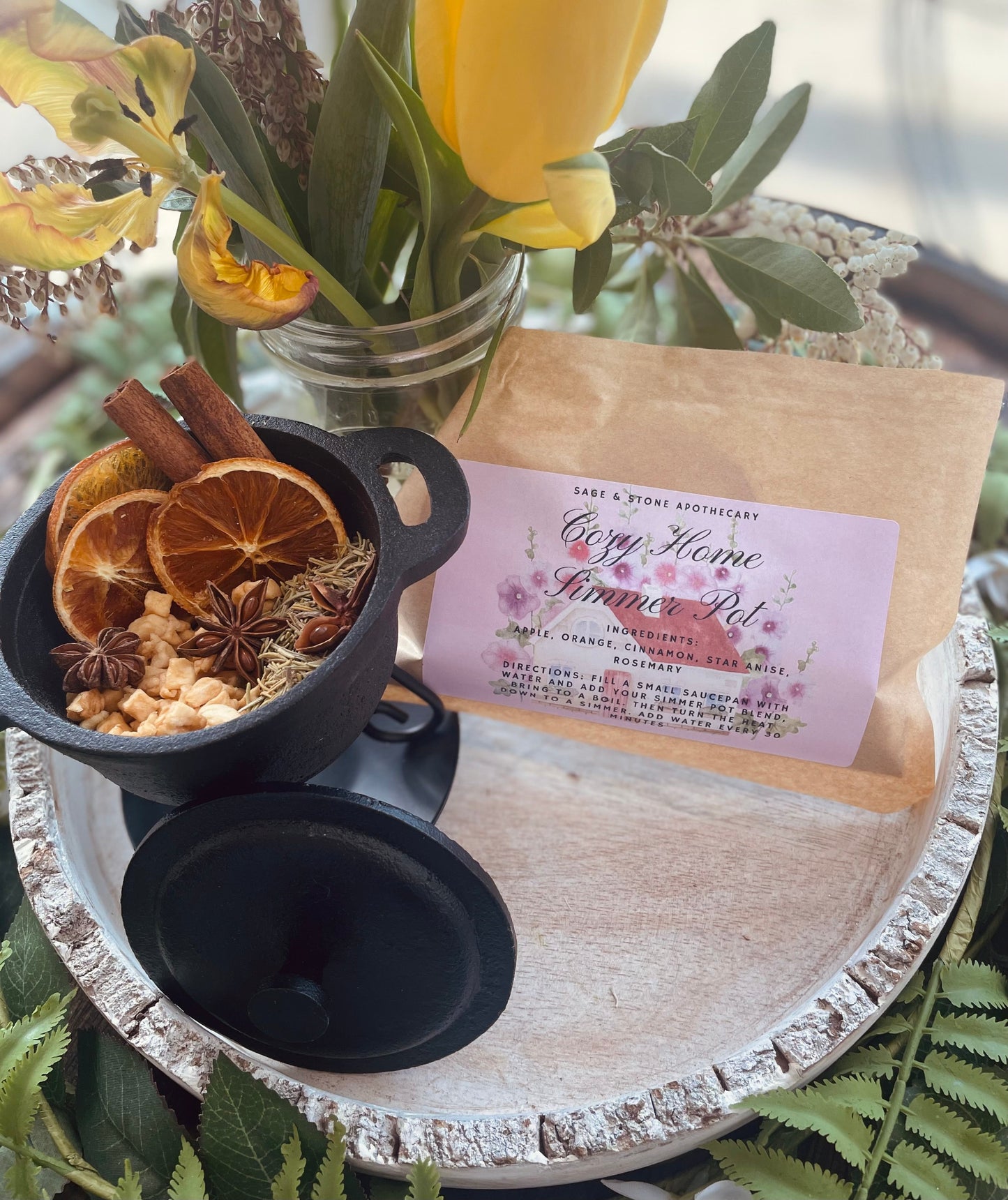 A brown kraft pouch with a pink floral label next to a small black pot filled with dried orange slices and cinnamon sticks, styled beautifully with fresh yellow tulips.
