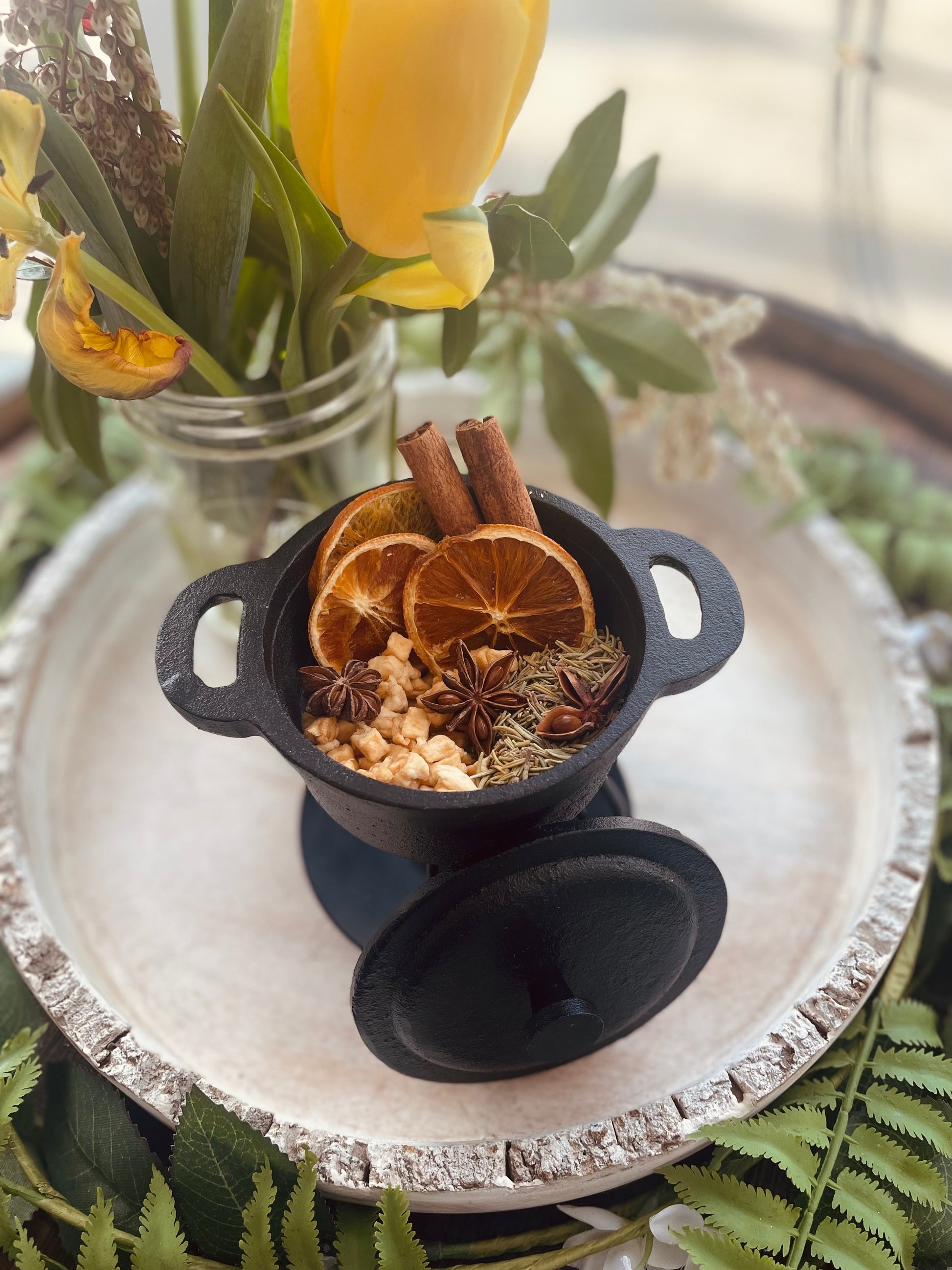 An overhead view of a black cast-iron style mini pot filled with aromatic dry spices and citrus slices resting on a white wooden decorative tray.