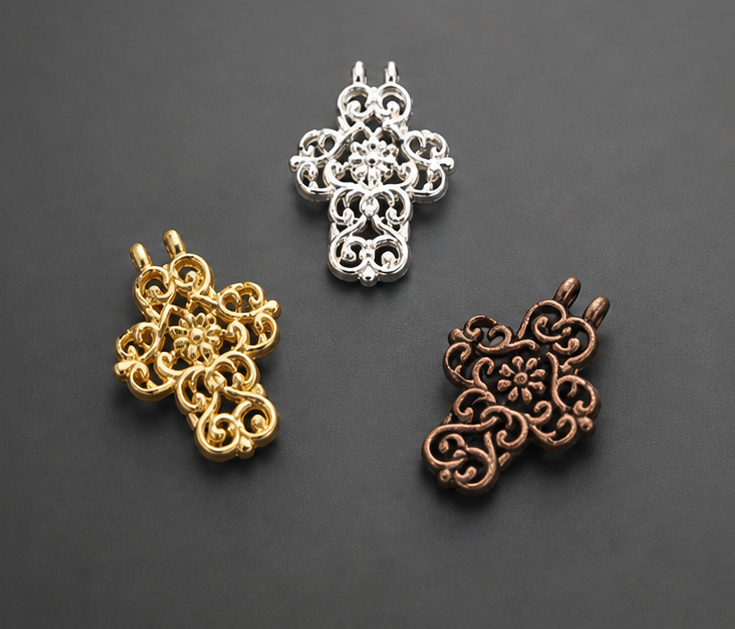 Three ornate filigree cross pendant charms showing the 22kt gold, silver, and antiqued copper metal options grouped together on a smooth dark gray surface.