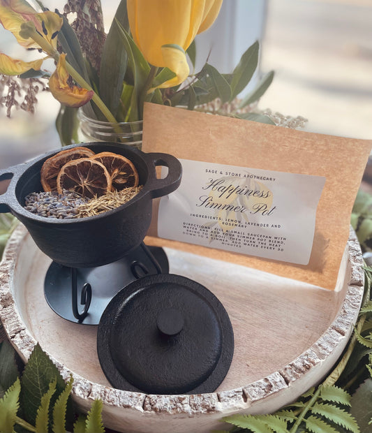 Close-up of the Happiness Simmer Pot Blend containing dried lemon slices, lavender, and rosemary inside a small black cast iron style cauldron pot next to its brown paper packaging.