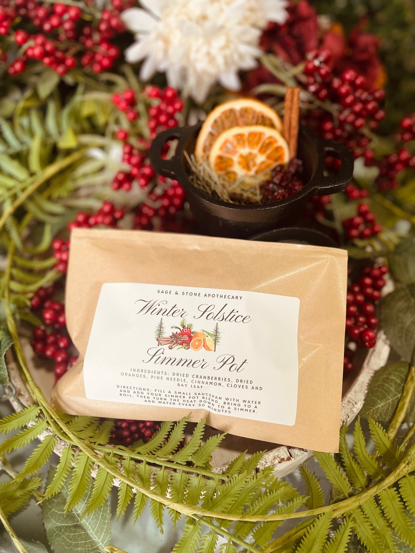 The eco-friendly Winter Solstice Simmer Pot package displayed sitting in front of a small black pot filled with the aromatic dried fruit ingredients.