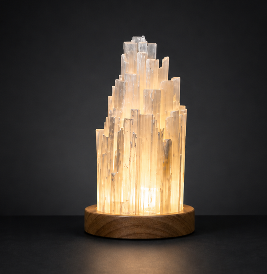 An ambient view of the illuminated raw selenite crystal skyscraper lamp casting a soft warm glow against a stark dark background.