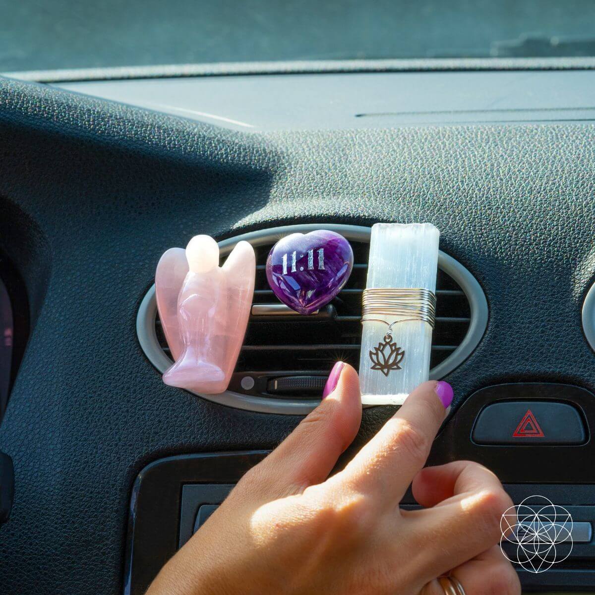 A close-up of a person's hand gently adjusting the selenite and amethyst crystal vent clips onto a car air conditioning vent in the sunlight.