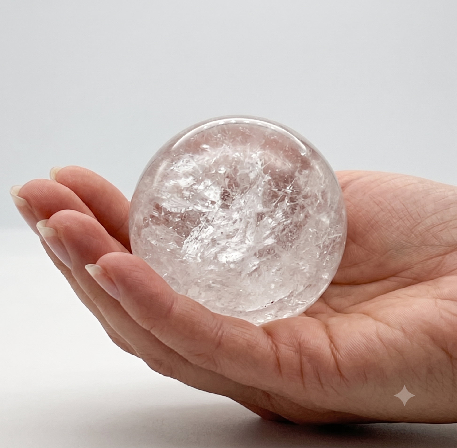 A hand cradling a highly clear, polished natural clear quartz crystal sphere displaying internal fracture patterns and light reflections.