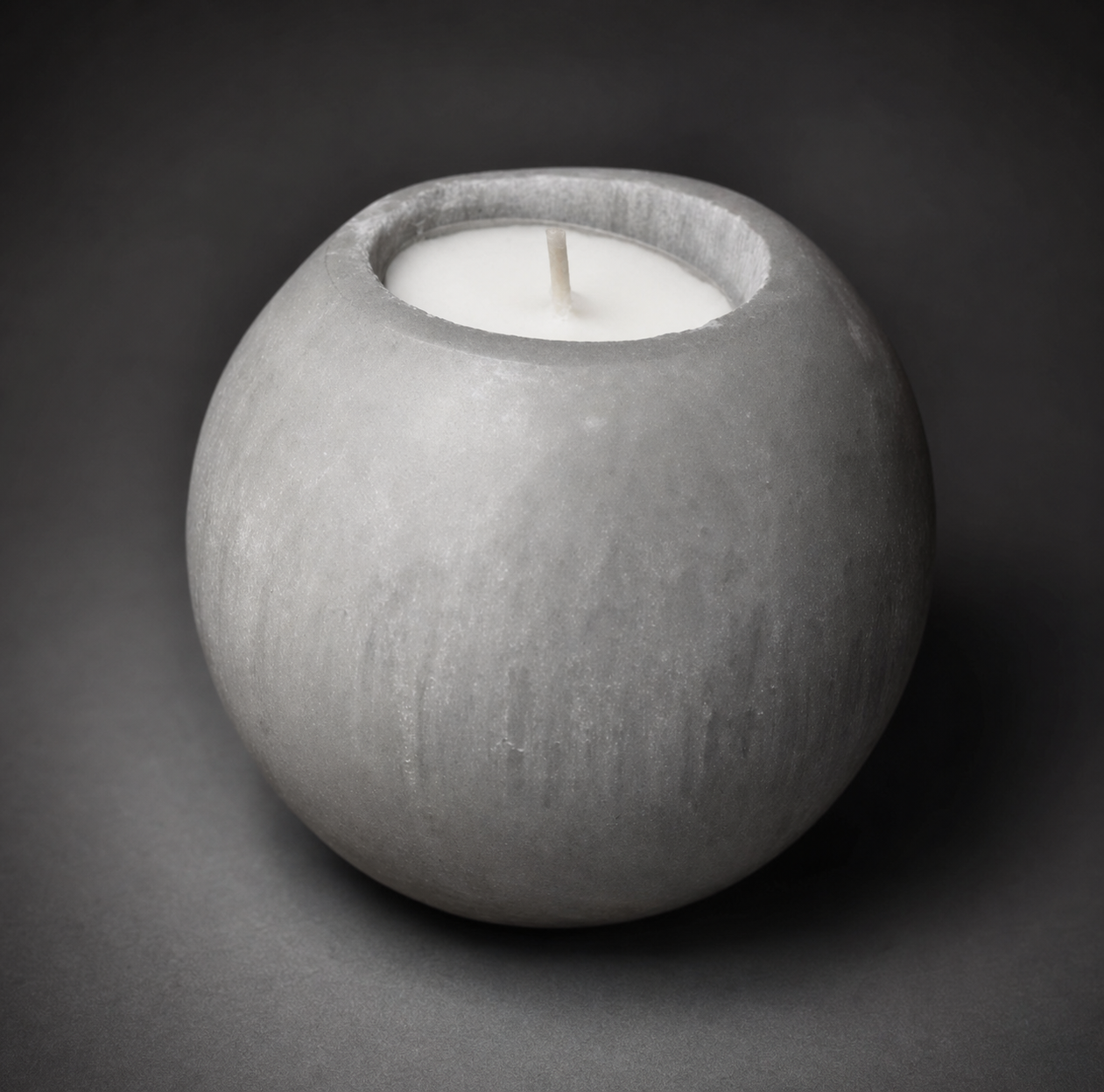 A hand-carved spherical white selenite crystal tealight candle holder with a lit wax candle inside, showing the soft glow through the natural stone fibers against a dark grey background.
