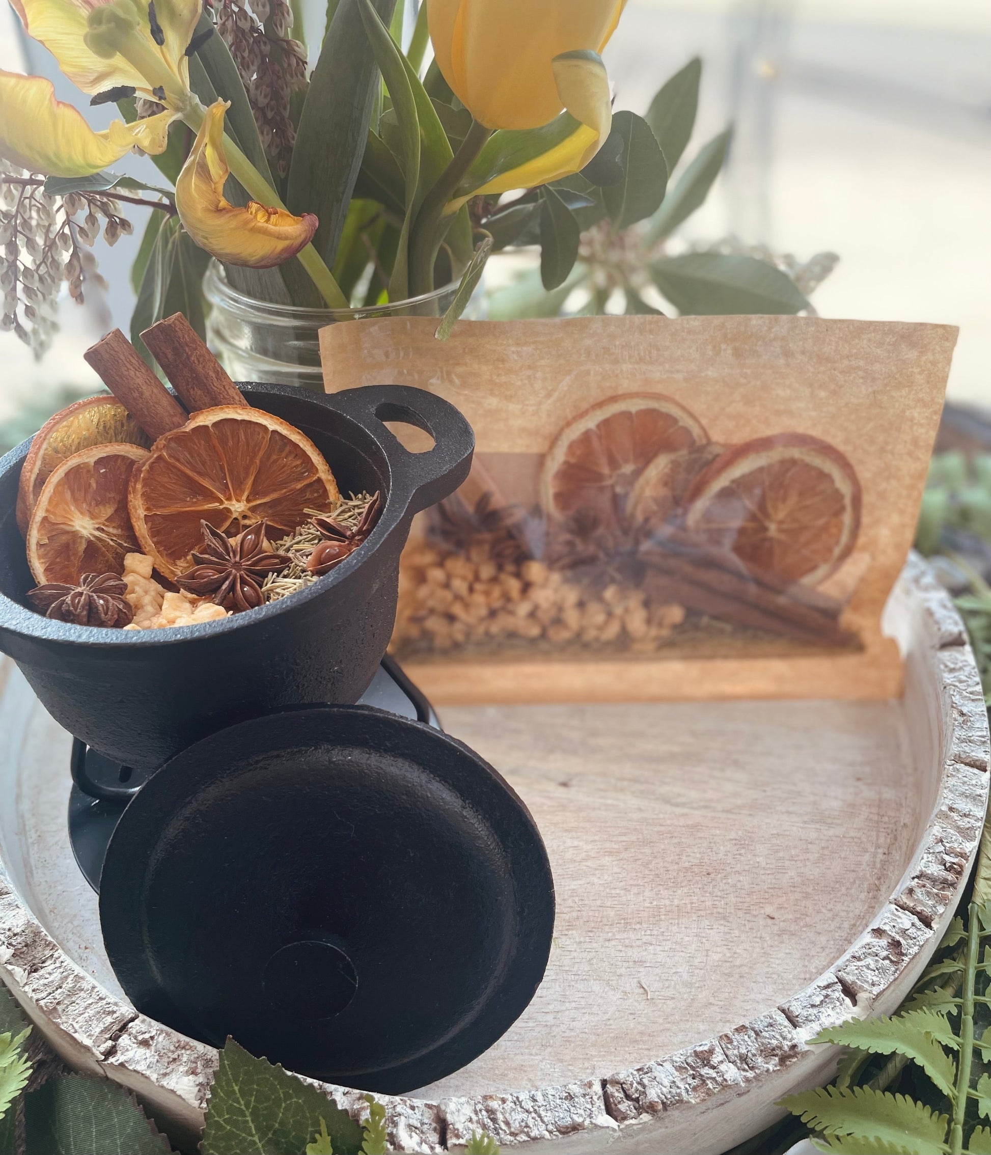 A small black simmer pot displaying dried orange slices and star anise next to a decorative wooden block art piece on a rustic round tray.