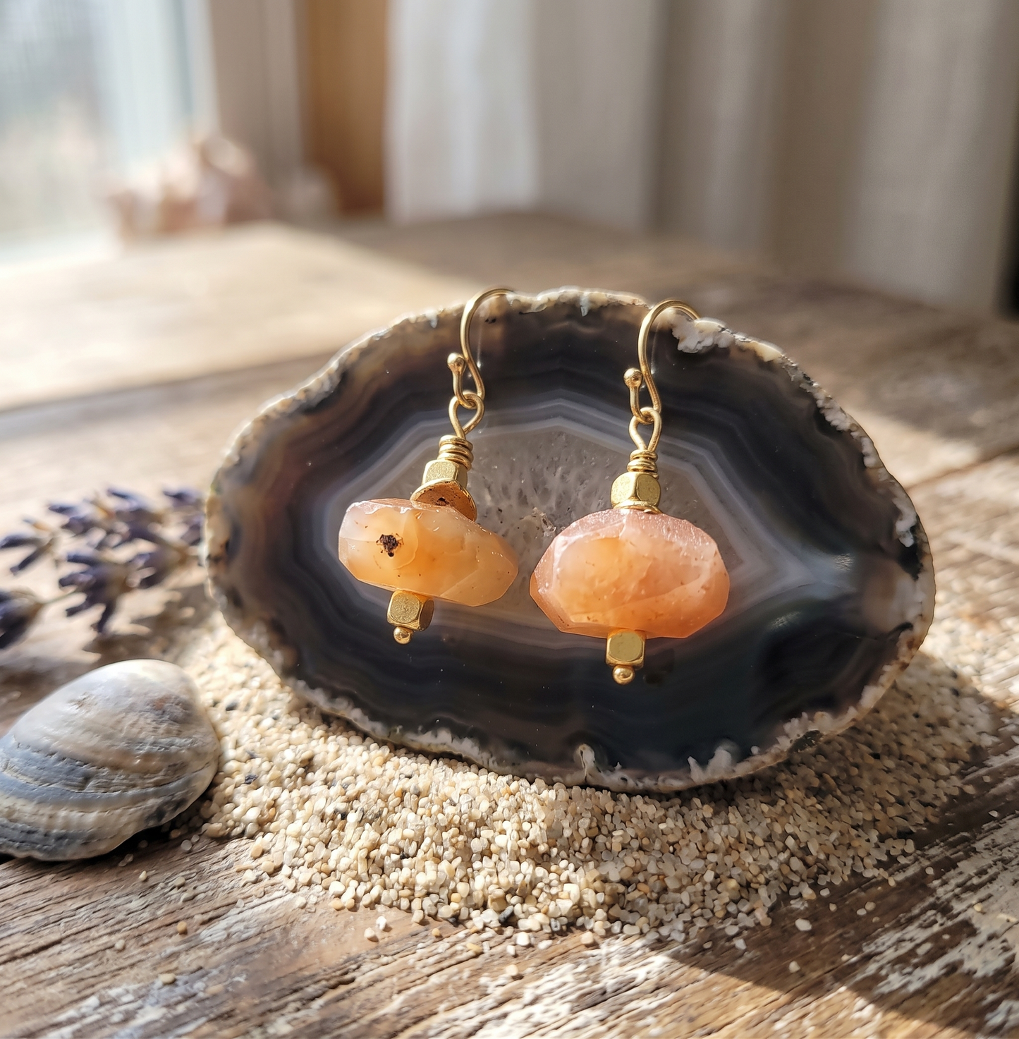 A pair of faceted orange agate gemstone earrings displayed on a dark crystal geode slice on a rustic wooden table.