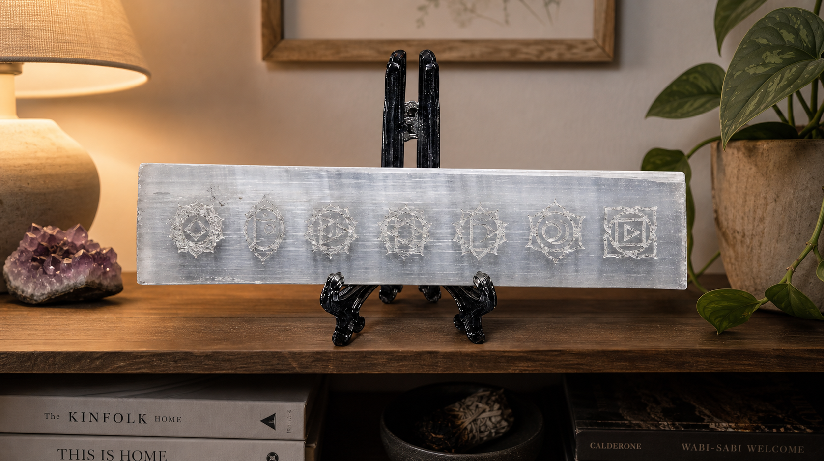 A long rectangular white selenite crystal plate engraved with seven intricate chakra symbols, displayed on a black ornate easel stand on a wooden tabletop with home decor accents.