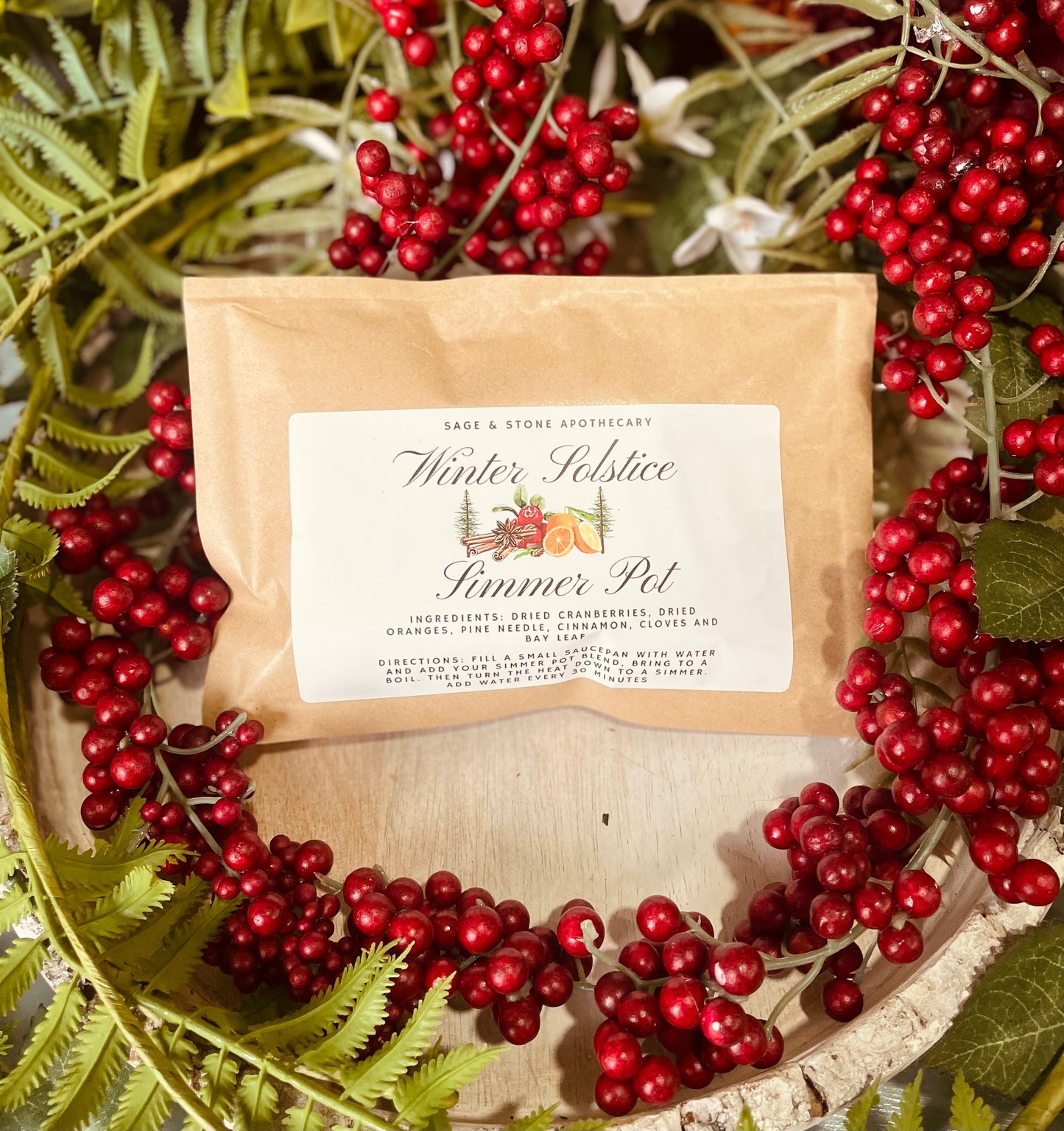 The front label of the brown kraft paper packaging pouch for the Winter Solstice Simmer Pot Kit resting on a festive bed of red winter berries.